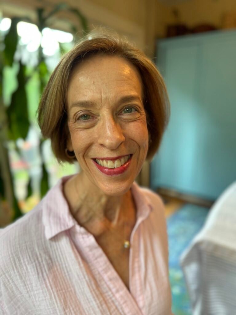 A woman with short brown hair, wearing a light pink blouse, smiles at the camera indoors with a blurred background.