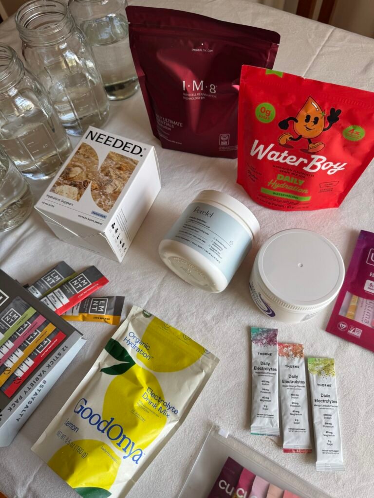 Various wellness and nutrition products, including supplements, snacks, drink mixes, and jars of water, are arranged on a white tablecloth.