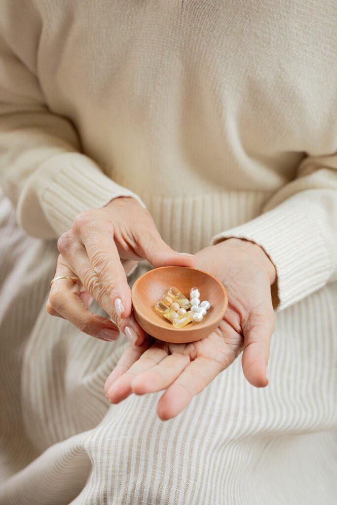 The 9 Best Longevity Supplements For Aging Well A person in a cream-colored outfit holds a small wooden bowl with assorted supplements in their hands.