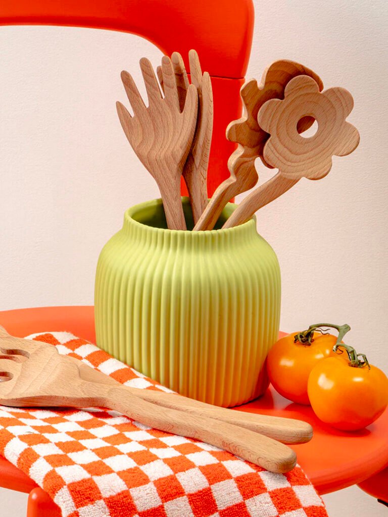 Wooden kitchen utensils in a green vase on an orange chair, with more utensils on an orange checkered cloth. Two orange tomatoes are next to the cloth.