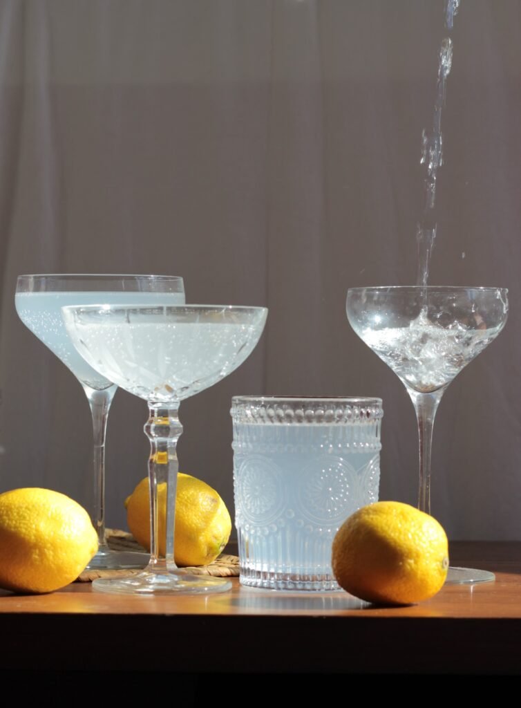 Three glasses filled with a cloudy beverage are surrounded by three whole lemons, with liquid being poured into one of the glasses, set on a dark wooden surface against a neutral background.