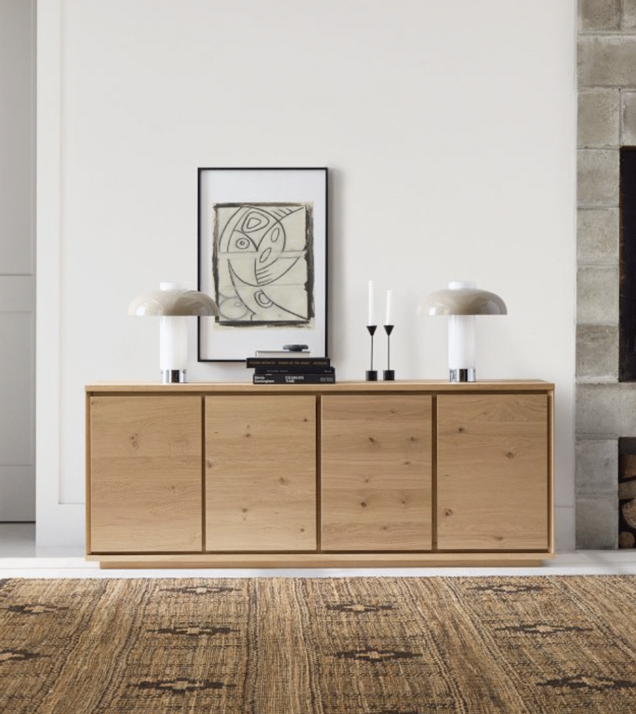 A wooden sideboard with two lamps, stacked books, and two candlesticks stands against a white wall with framed abstract art above, next to a stone fireplace.