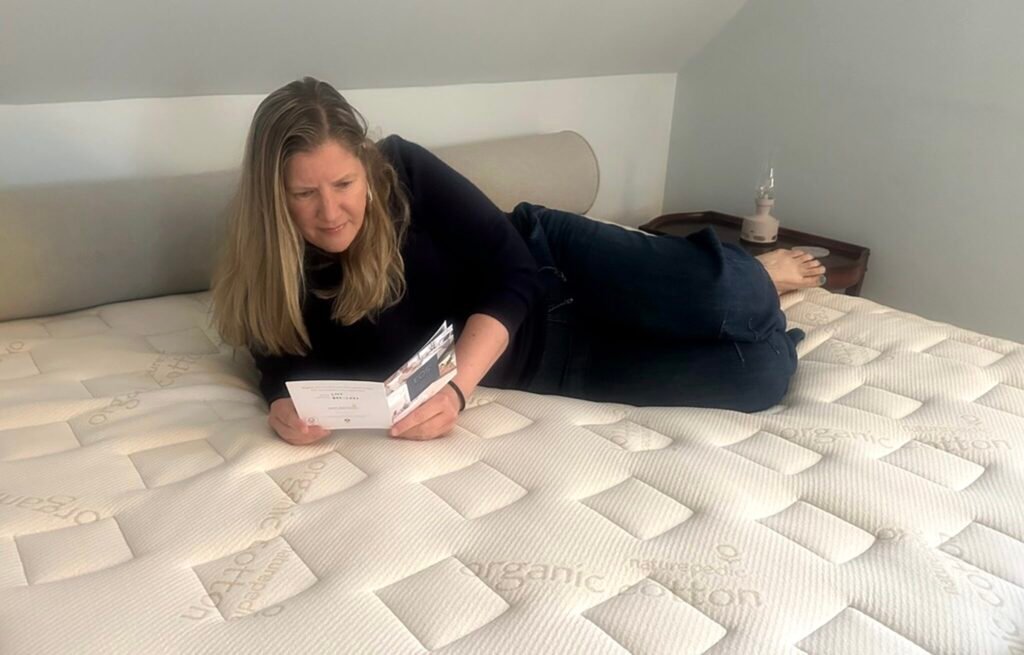 A Holistic Therapist Reviews Naturepedic’s EOS Mattress A woman lies on a bed reading a pamphlet. The mattress surface is labeled "organic cotton." A nightstand with a lamp is nearby.