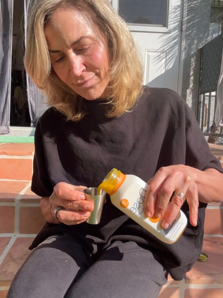 A woman sits on outdoor steps, pouring a yellow beverage from a plastic bottle into a small metal cup.