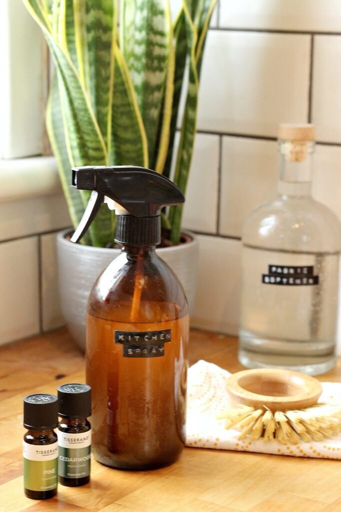 Amber glass bottle full of DIY cleaning spray in a white metro tiled kitchen, next to essential oils and natural cleaning supplies.