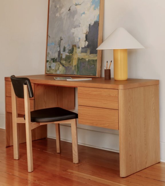 9 Best Sustainable Wood Desks For 2026 A wooden desk with four drawers holds a lamp, a painting, pencils in a cup, and a closed laptop. A wooden chair with a black seat is placed in front of the desk.