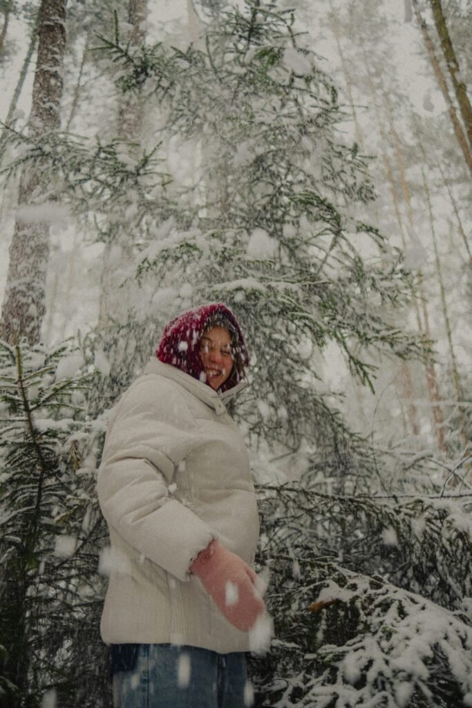 Using The Wheel Of The Year For A Deeper Connection To The Seasons A person in a white winter coat and red hat stands in a snowy forest as large snowflakes fall around them.
