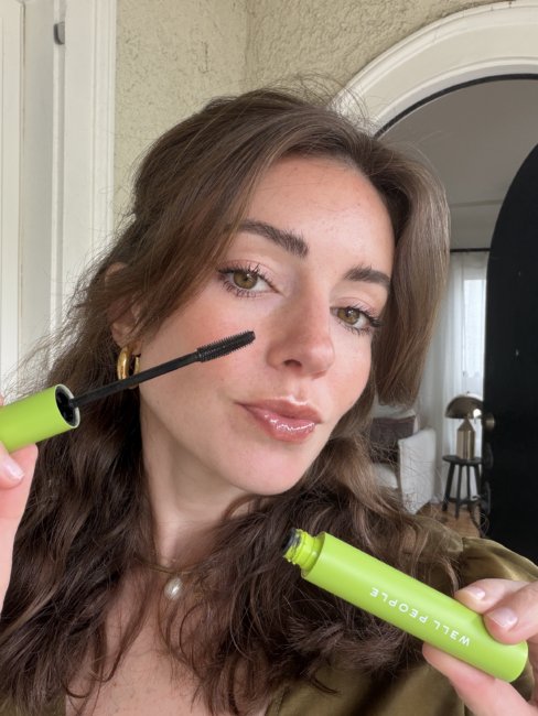 We Review Well People’s Expressionist Curling Mascara (2026) A woman with wavy brown hair applies mascara to her eyelashes using a wand. She is holding a green tube labeled “WELL PEOPLE.”.
