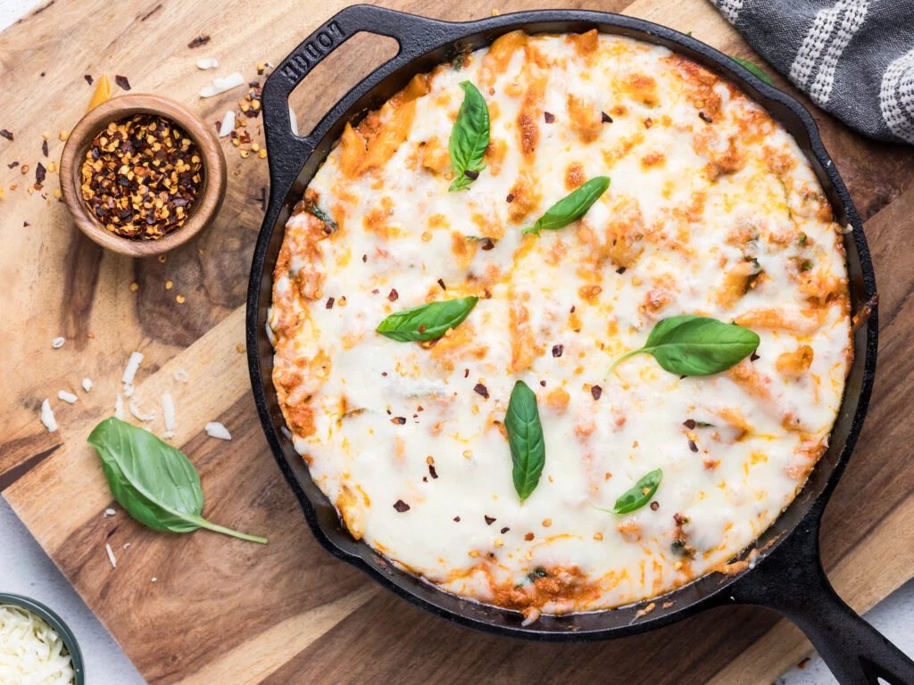 10 Healthy Dinners in 30 Minutes or Less A cast iron skillet filled with baked cheesy pasta topped with melted cheese, fresh basil leaves, and sprinkled red pepper flakes. The skillet is on a wooden board with a small bowl of red pepper flakes and basil nearby.