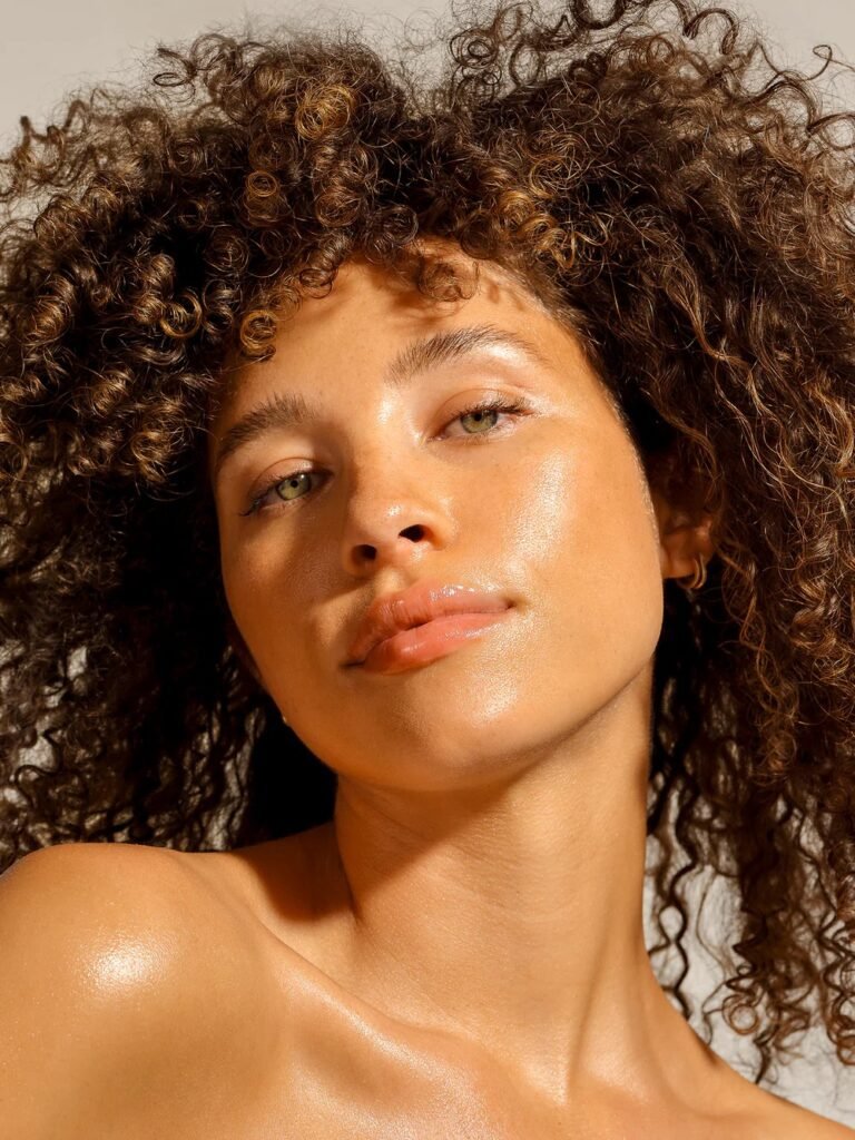 9 Nontoxic Self Tanners For A Healthy Glow (2026) A woman with curly hair and glowing skin looks slightly upward with a neutral expression against a plain background.