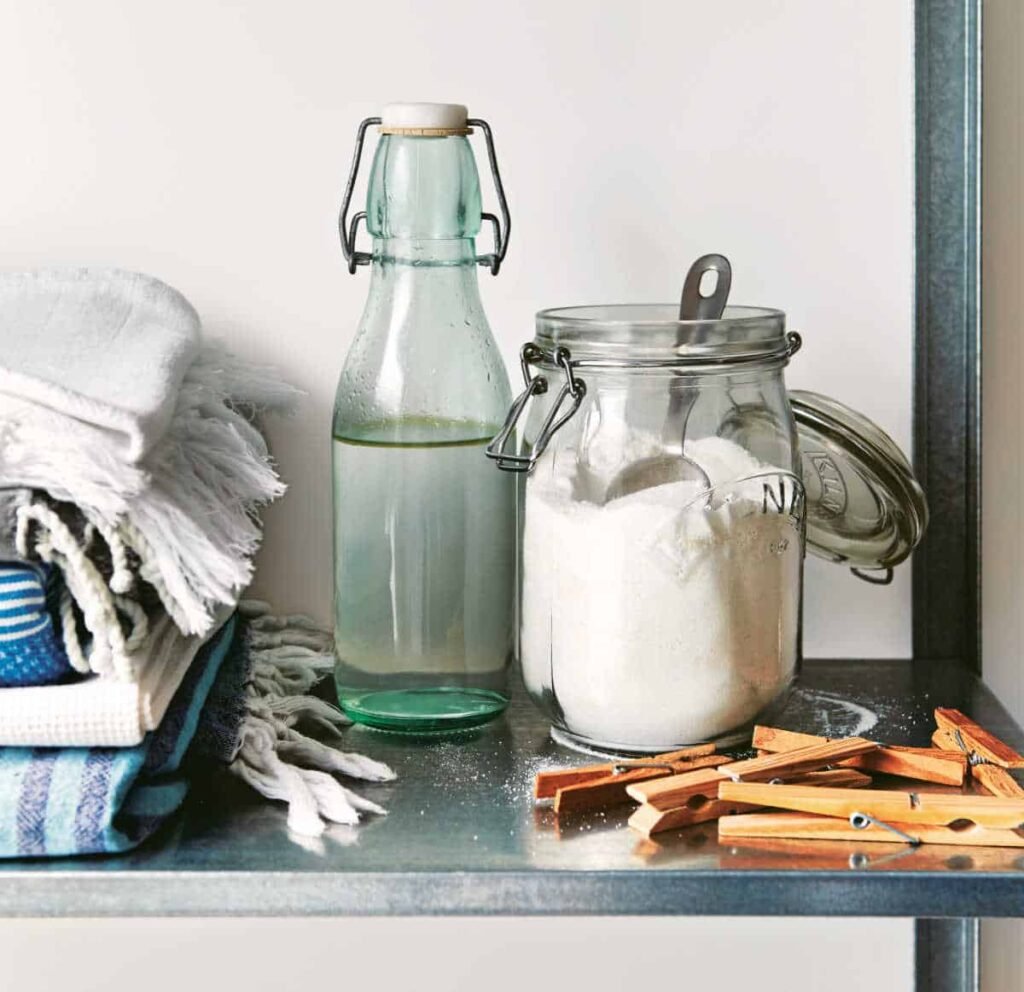 Jar of soda crystals, next to a bottle of vinegar, some folded laundry and some wooden pegs