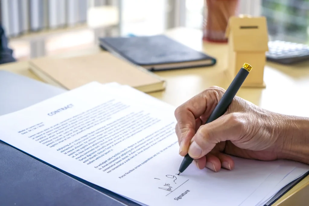 8 Best Term Life Insurance Companies A persons hand holding a pen signs a contract document on a desk, with other paperwork, a notebook, and a small wooden box in the background.