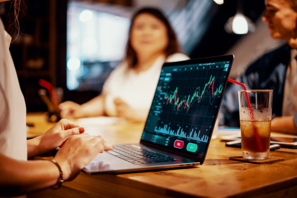 Index Funds Beat Most Actively Managed Funds Over Time A person uses a laptop displaying a colorful stock market chart in a meeting setting, with two blurred people and a drink with a straw in the background.